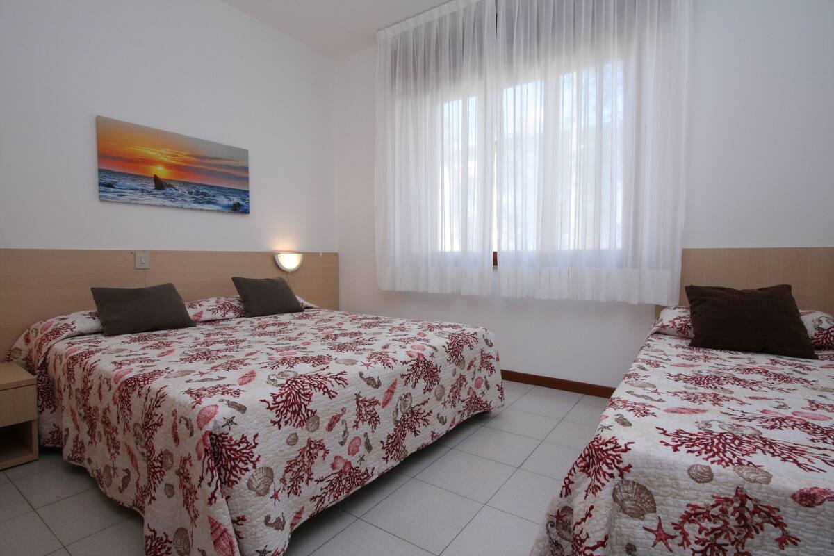 A hotel room with two beds, a painting of a sunset over the ocean, and a large window with sheer curtains.