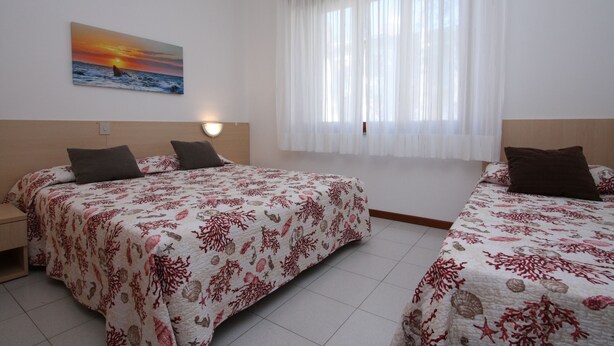 A hotel room with two beds, a painting of a sunset over the ocean, and a large window with sheer curtains.