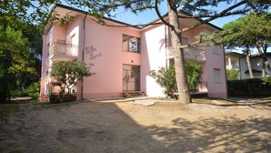 A pink building with a balcony and a door, surrounded by trees and a gravel driveway.