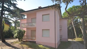 A two-story pink building with a balcony on the second floor, surrounded by trees and a gravel path.