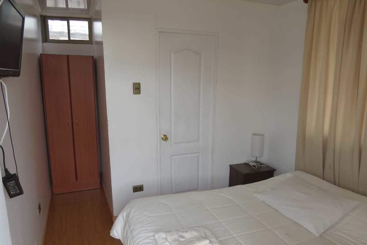 Ein Hotelzimmer mit Bett, Kleiderschrank, Tür, Nachttisch mit Lampe und einem an der Wand montierten Fernseher.