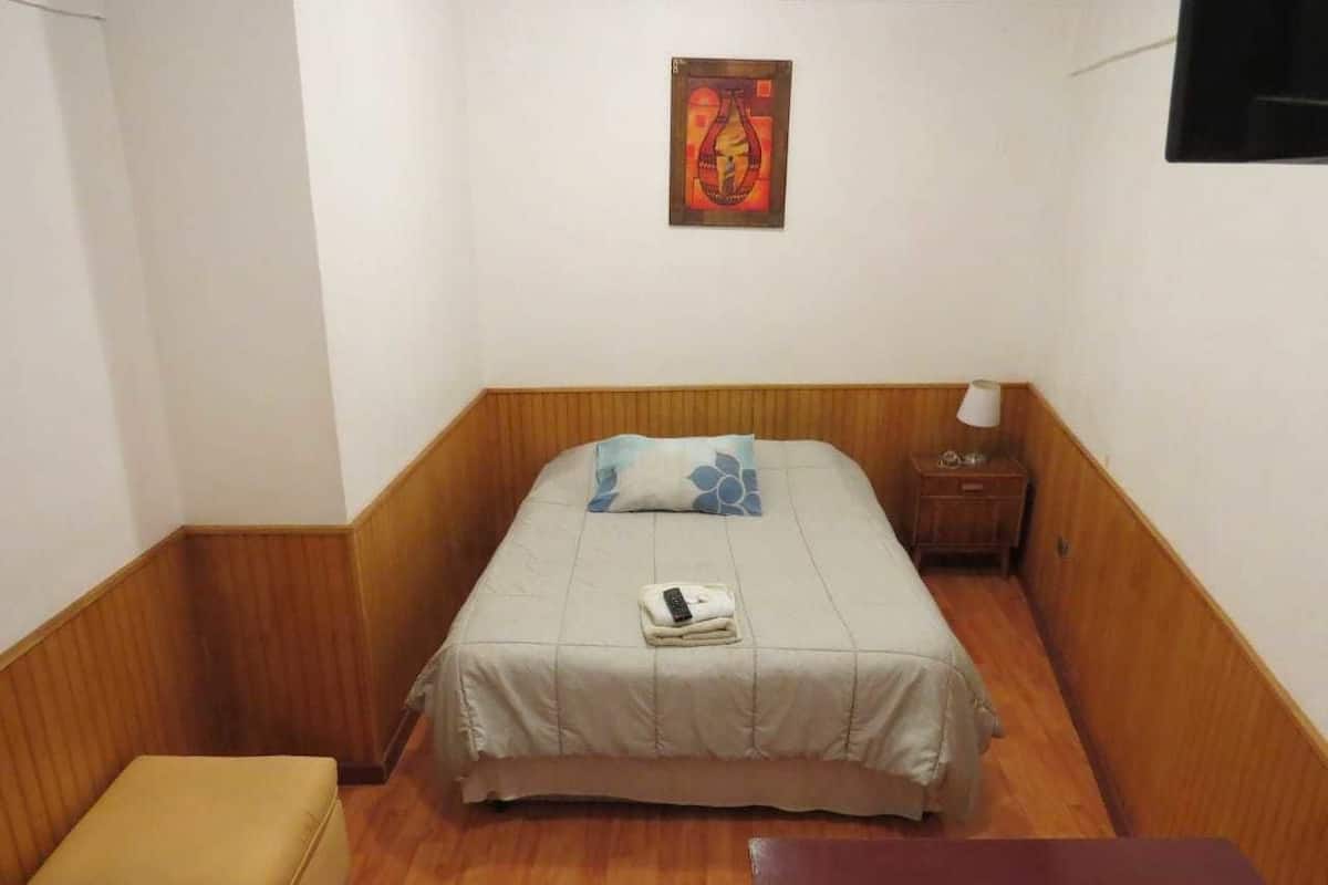 Ein kleines Hotelzimmer mit einem Bett, einem Nachttisch, einer Bank und einem Wandbild.