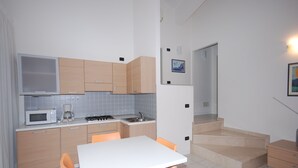 Een kleine keuken met een magnetron, fornuis en wastafel. Er is een eettafel met stoelen en een trap naar een hoger gelegen verdieping.