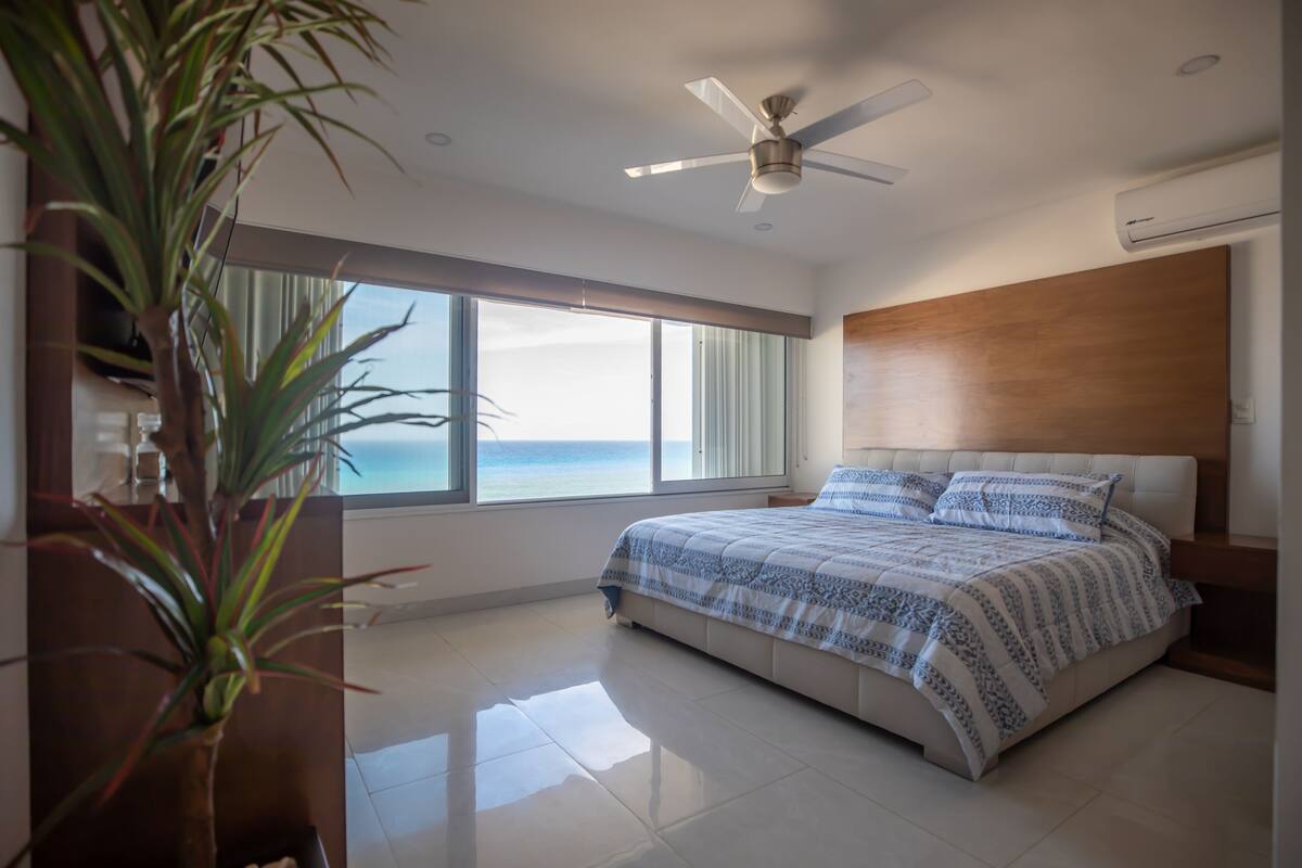 A modern bedroom with a large bed, a ceiling fan, and a view of the ocean.