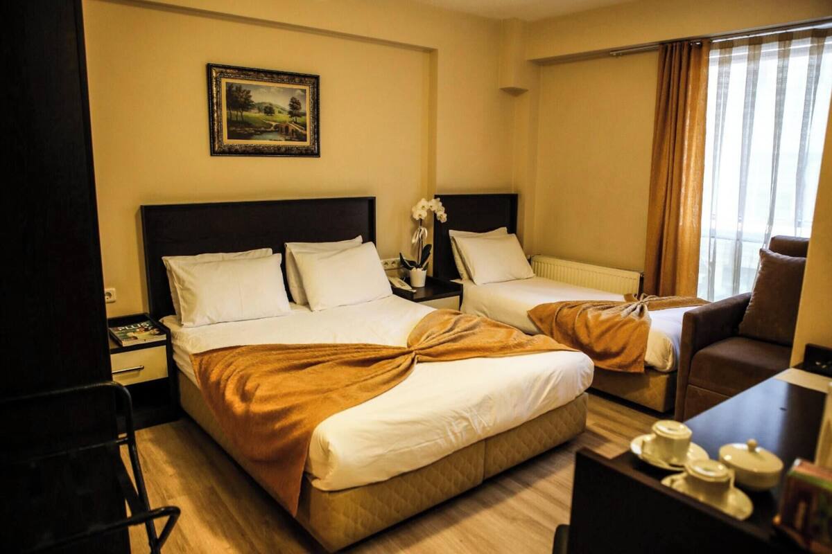 A hotel room with two beds, a desk, a chair, a small table with cups, and a window with curtains.