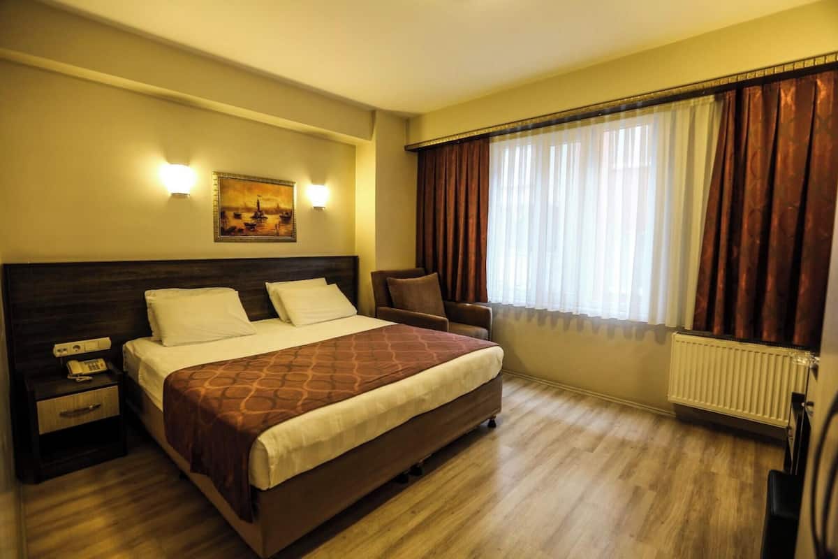 A hotel room with a bed, two bedside tables, a telephone, a painting on the wall, and a window with curtains.