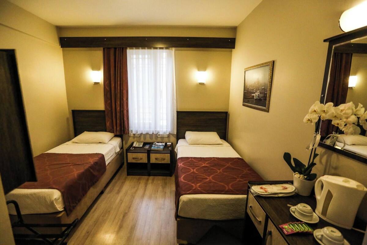 A hotel room with two beds, a desk with a telephone and books, a mirror, and a window with curtains.