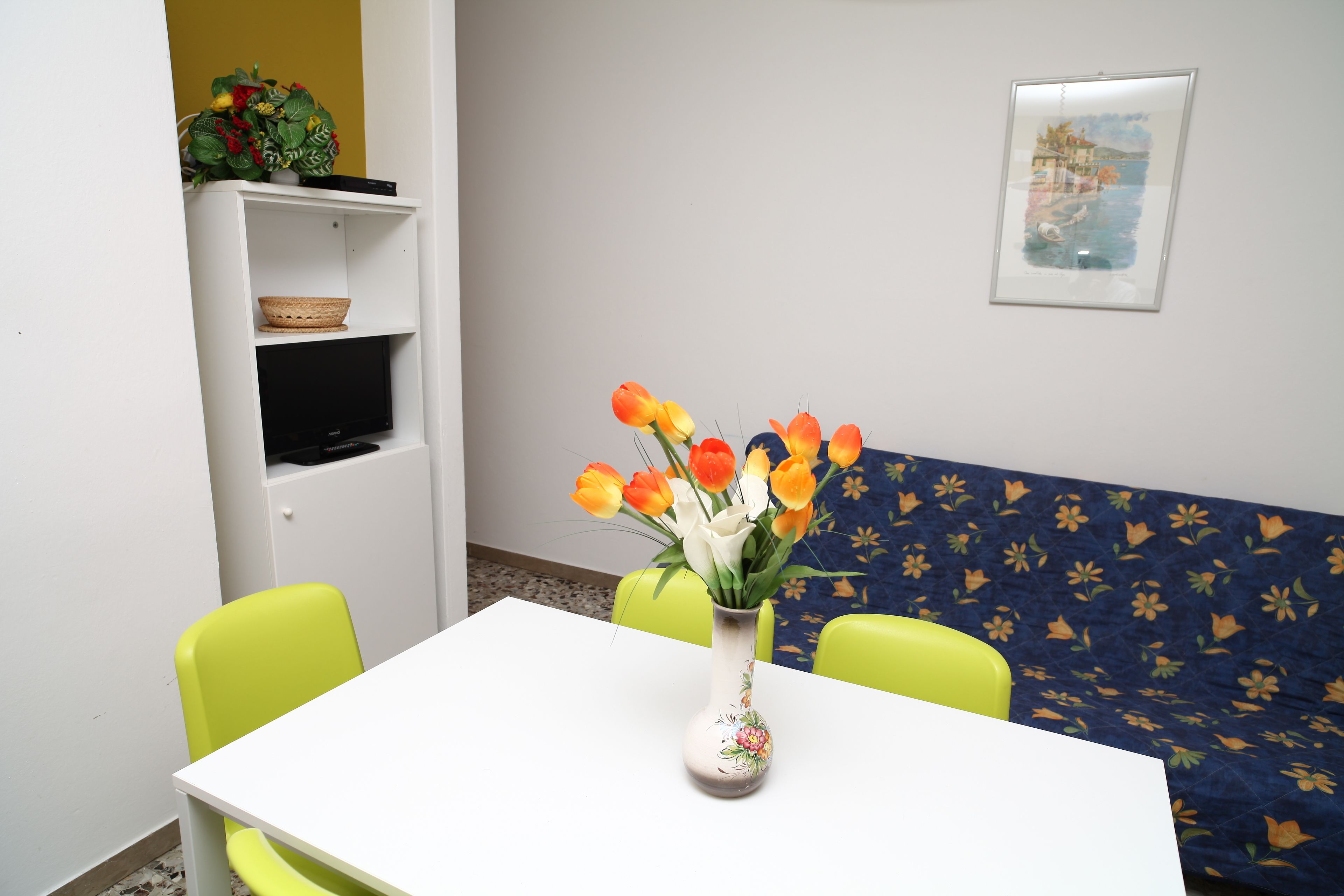 A room with a white table, a vase with flowers, a green chair, a sofa with a floral pattern, and a shelf with a basket and a plant.