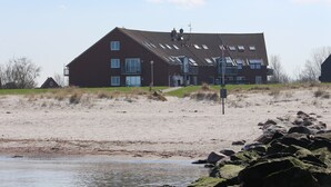 Suuri, kaksikerroksinen talo, jossa on useita ikkunoita ja kalteva katto. Se sijaitsee hiekkarannalla, ja sen edessä on kivikkoinen rantaviiva.