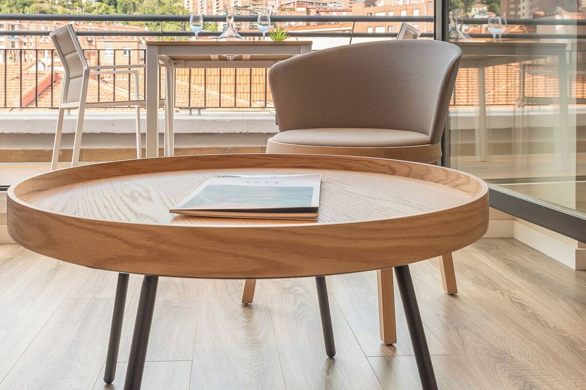 Una habitación moderna con una mesa redonda de madera y dos sillas, un ventanal con vistas a un paisaje urbano y zonas verdes.
