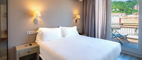 Habitación de hotel con una cama grande, dos lámparas de noche, una mesita de noche y vistas a un paisaje urbano desde un balcón.