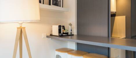 A modern kitchen with a bar area, white bar stools, a floor lamp, and framed pictures on the wall.