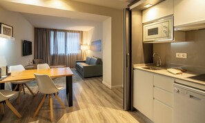 Une chambre d’hôtel moderne avec une table à manger en bois, des chaises blanches, un canapé et une kitchenette équipée d’un four à micro-ondes et d’un évier.
