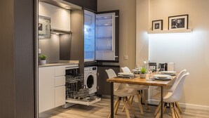 A compact kitchen with a dishwasher, a built-in oven, and a dining area with a wooden table and white chairs. - Bilbao City Center by abba Suites (Bilbao)