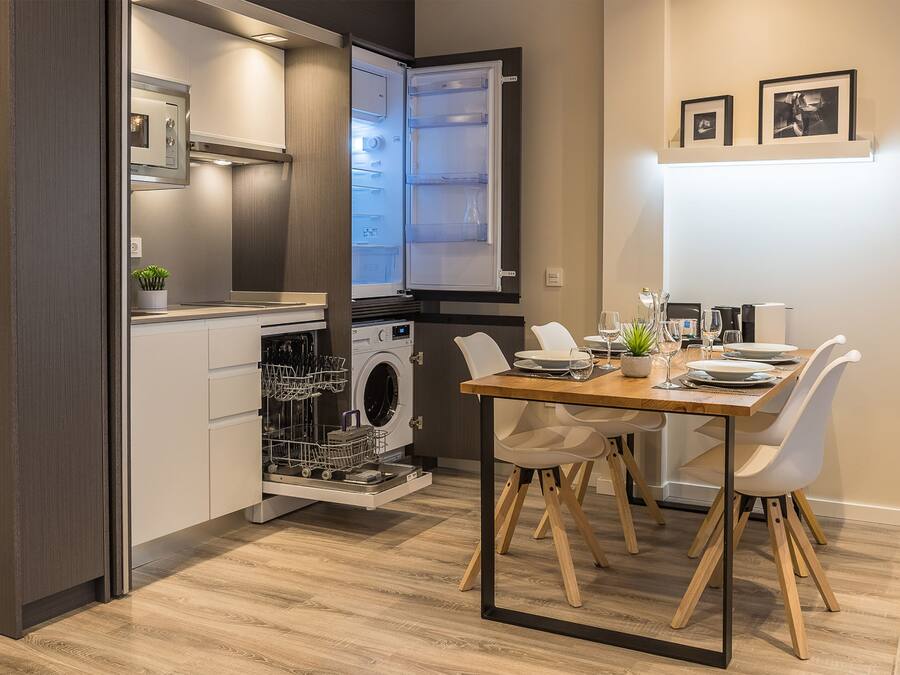 Cocina compacta con lavavajillas, horno empotrado y comedor con mesa de madera y sillas blancas.