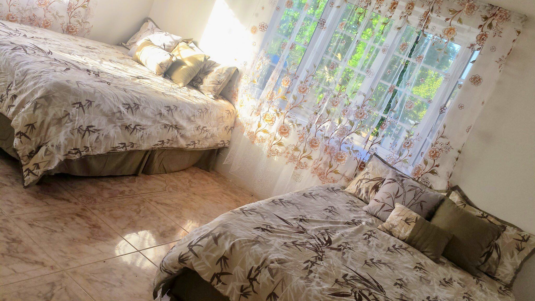 A bedroom with two beds, floral-patterned bedding, a window with floral curtains, and a tiled floor.