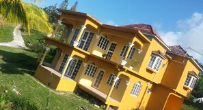 A multi-story yellow building with multiple balconies, a red roof, and a pathway leading to the entrance.