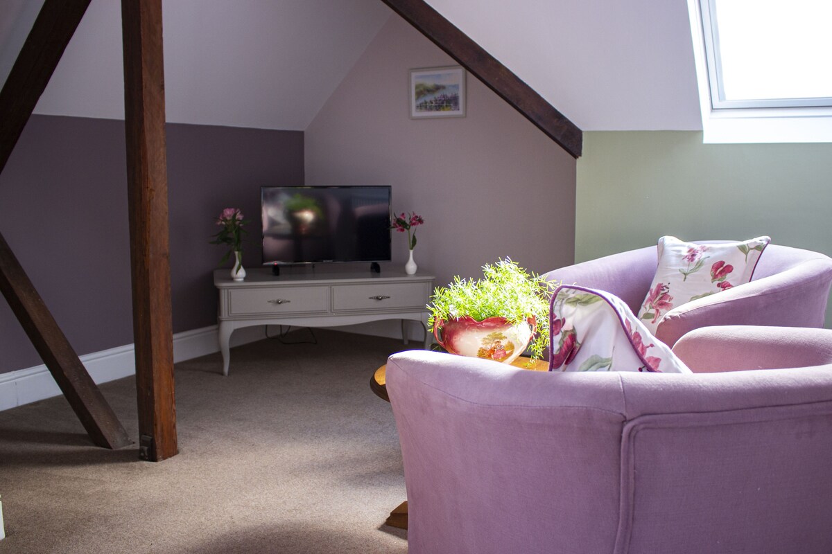 A room with a sloped ceiling, a television on a white dresser, a purple armchair, and a potted plant.