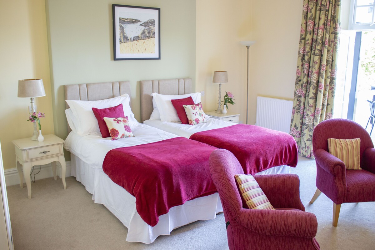 A hotel room with two beds, a red armchair, a nightstand with a lamp, and a framed picture on the wall.