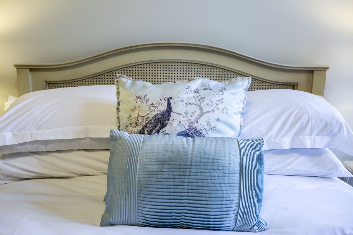 A bed with white linens, a decorative pillow with a peacock design, and a blue striped pillow.
