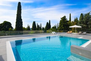 A rectangular swimming pool with clear blue water, surrounded by a glass fence. There are lounge chairs and a white umbrella on the poolside. - Minnie (Gardone Riviera)