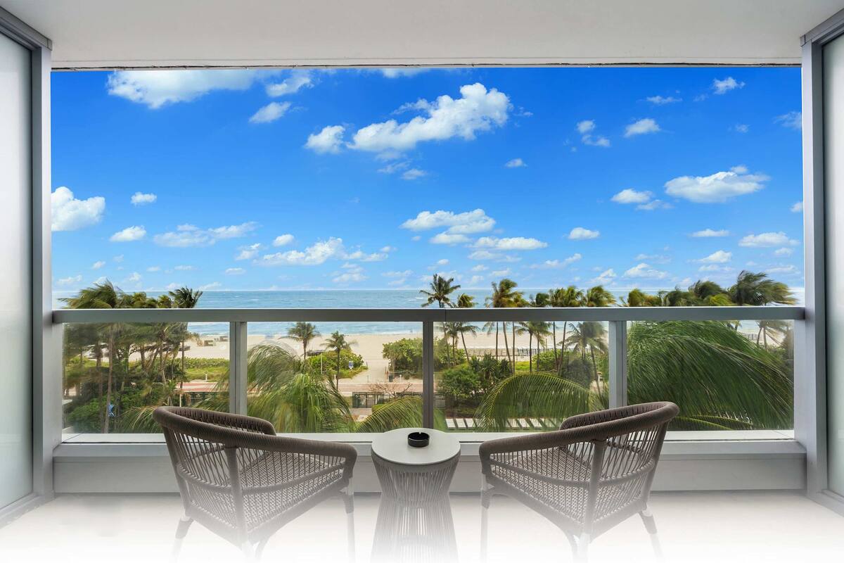 Een balkon met rieten stoelen en een kleine tafel, met uitzicht op een strand met palmbomen en een helderblauwe lucht.