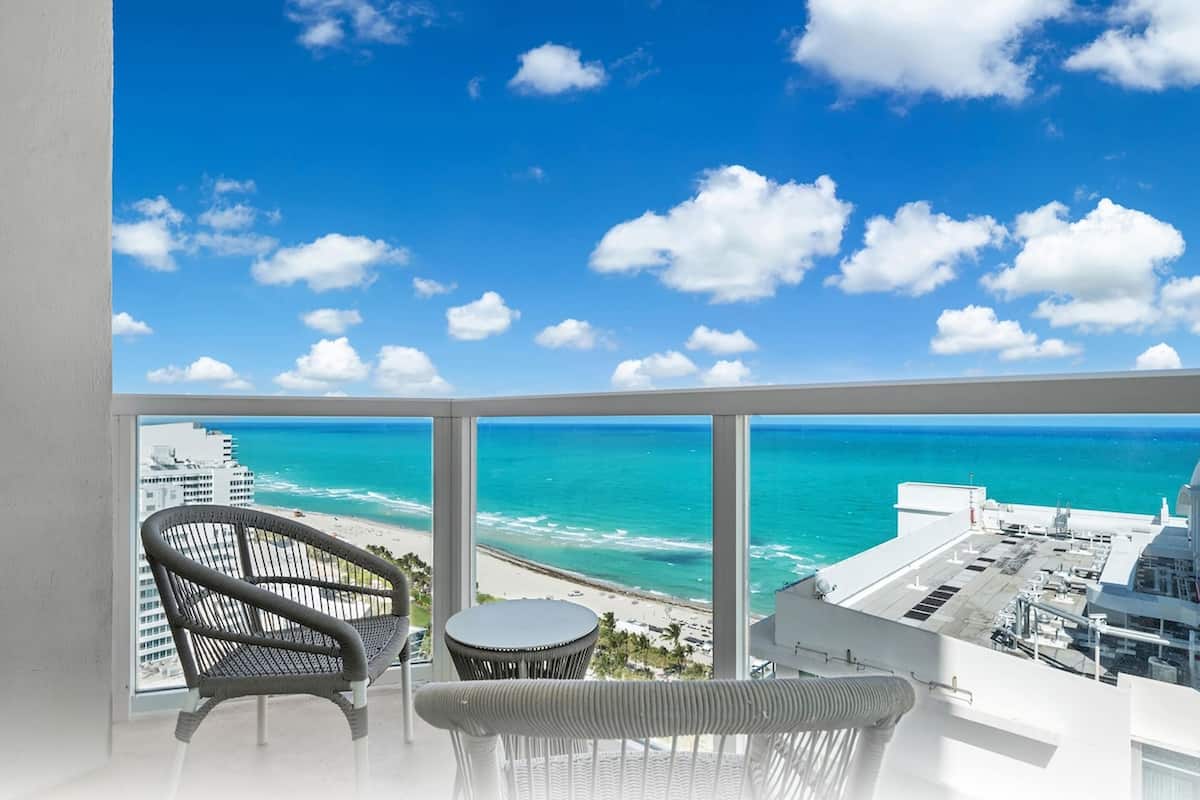 Een balkon met uitzicht op de oceaan en het strand, met twee rieten stoelen en een kleine ronde tafel.