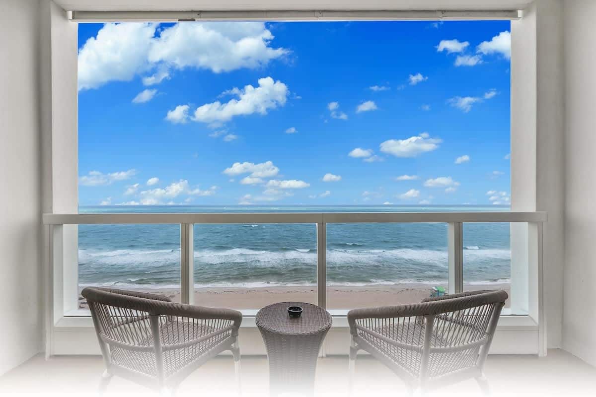 Een balkon met rieten stoelen en een kleintje tafeltje, met uitzicht op een strand met golven en een heldere lucht.