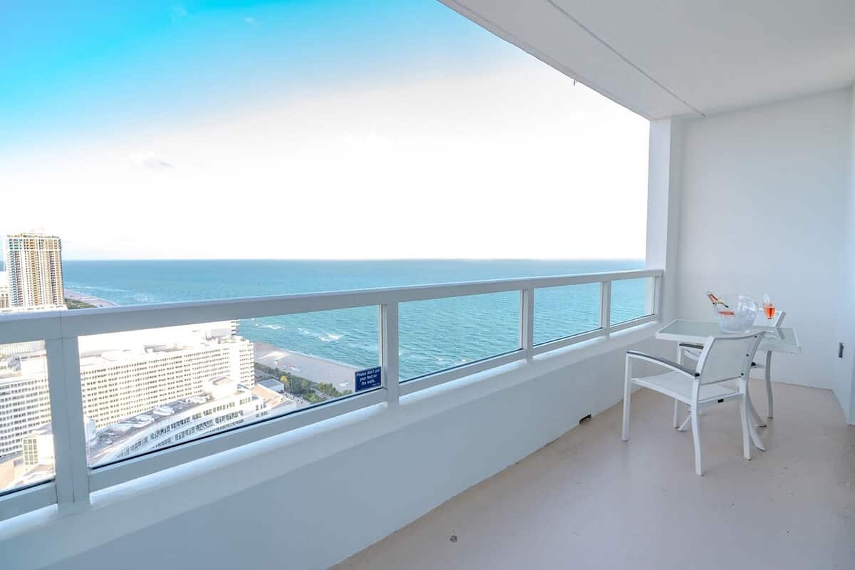 Een balkon met uitzicht op zee en stadsbeeld, een witte tafel met stoel, en een klein raam met een bord.