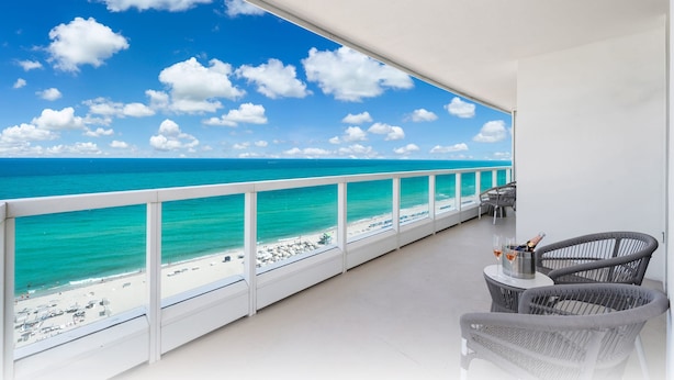 Een balkon met uitzicht op het strand en de oceaan. Er staan twee rieten stoelen en een klein tafeltje met een fles en glazen.