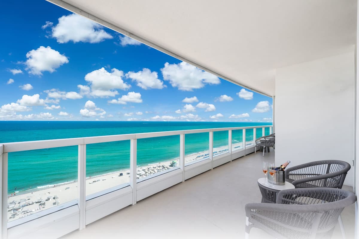 Een balkon met uitzicht op het strand en de oceaan. Er staan twee rieten stoelen en een klein tafeltje met een fles en glazen.