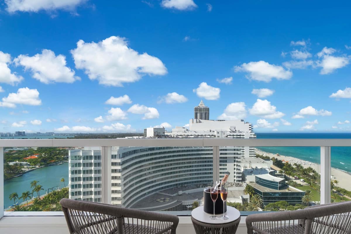 Een balkon met rieten stoelen, een tafel met fles en glas, met uitzicht op een strand en een stadsbeeld met een opvallend gebouw.