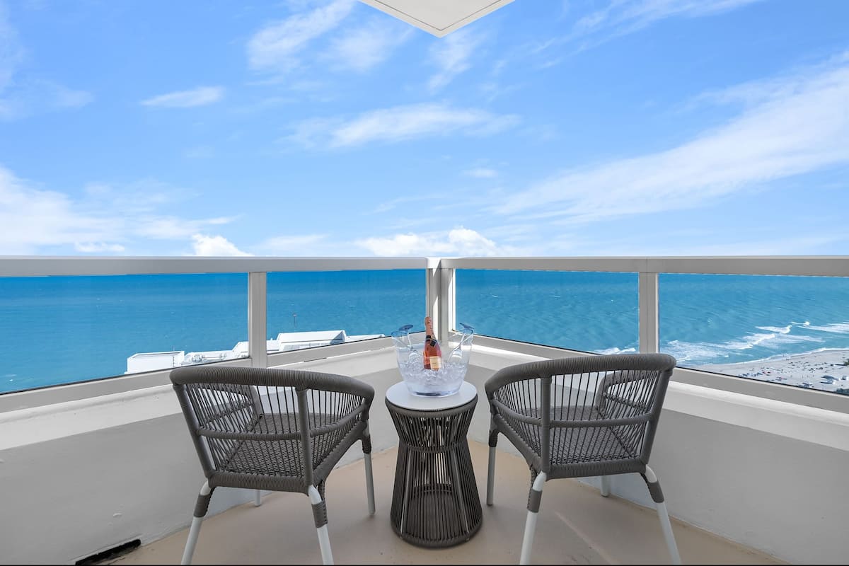 Een balkon met twee rieten stoelen, een klein rond tafeltje en een fles champagne. Uitzicht op de oceaan en een strand.