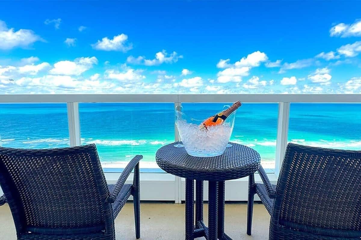 Een balkon met twee rieten stoelen, een klein rond tafeltje en een fles champagne in een glazen schaal. In de verte een heldere blauwe oceaan en een witte balustrade.