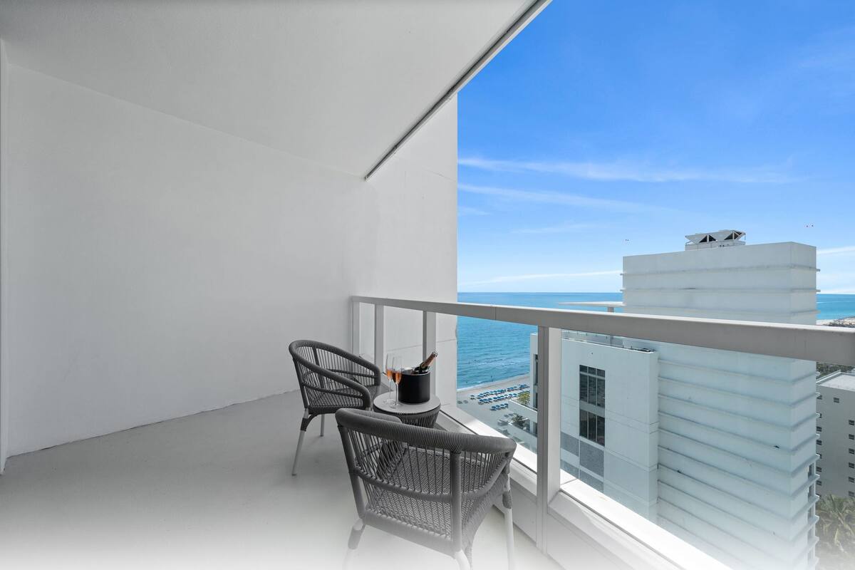 Een balkon met twee rieten stoelen, een klein tafeltje en uitzicht op zee en gebouwen.