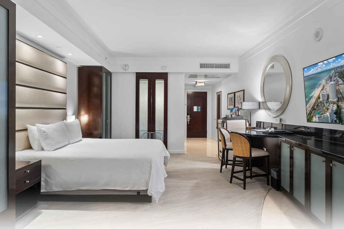 A modern hotel room with a large bed, a desk with a chair, a large mirror, and a painting of a coastal cityscape.