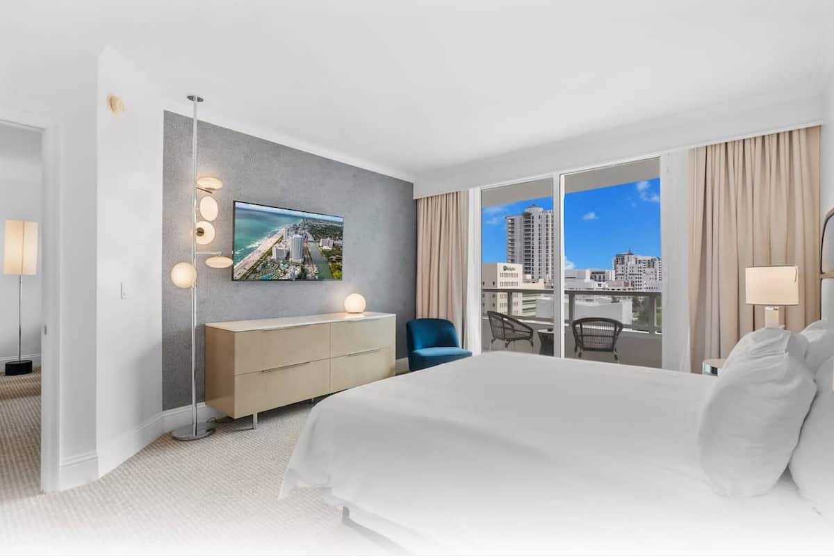 Een moderne hotelkamer met een groot bed, een flatscreen televisie, een dressoir en uitzicht op de stad door de balkondeuren.
