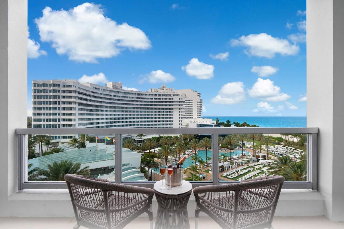 Een balkon met rieten stoelen en een tafel, met uitzicht op een resort met een zwembad en een groot hotelgebouw.