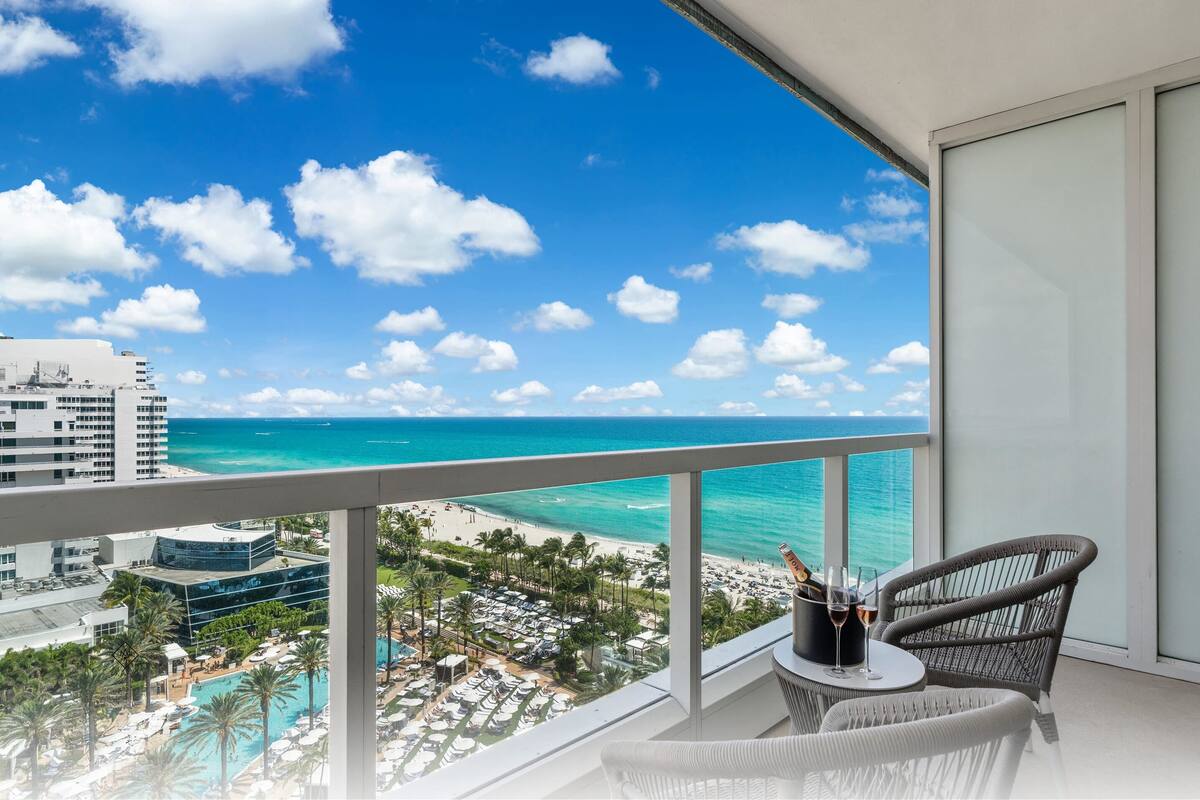 Een balkon met uitzicht op zee en strand, een zwembad en een eethoek met een stoel en tafel.