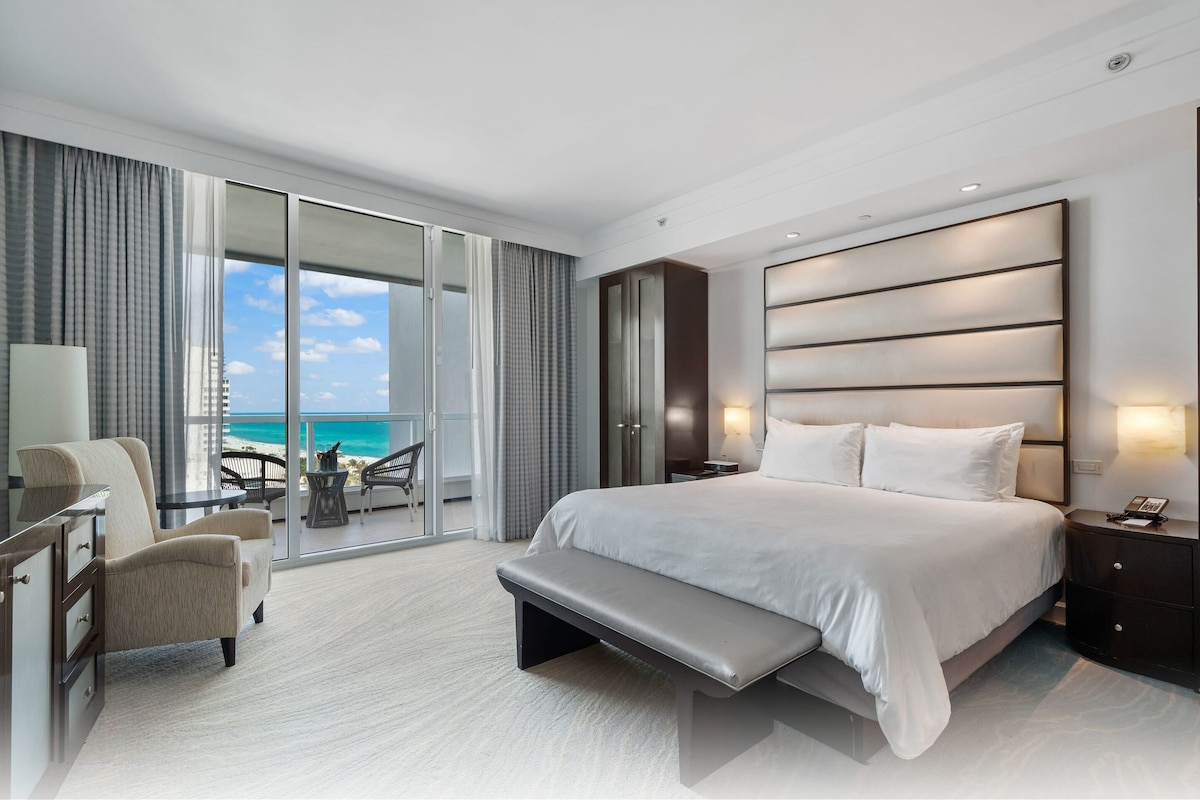 Een moderne hotelkamer met een groot bed, een bankje, een bureau en uitzicht op de oceaan.