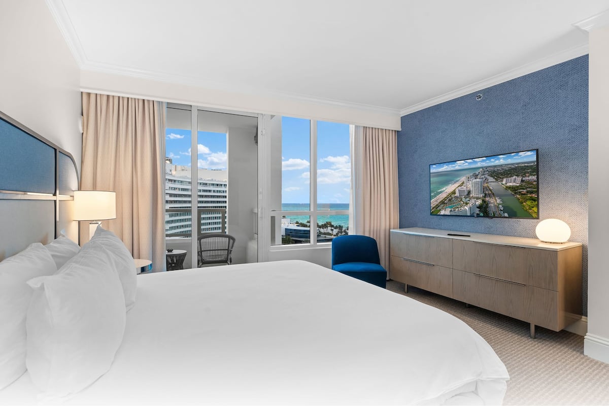 Een hotelkamer met een groot bed, een aan de muur bevestigde televisie, een bureau met een stoel, en uitzicht op de stad door het raam.