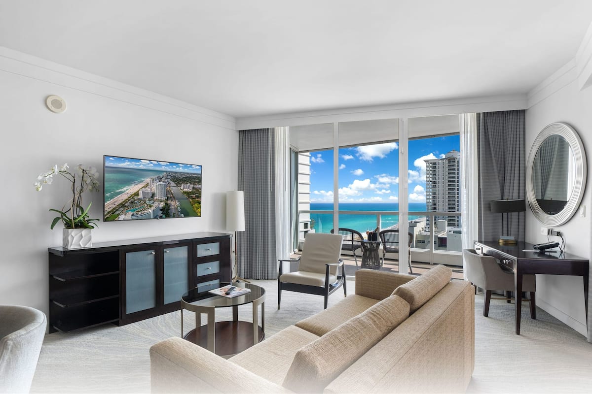 Een moderne hotelkamer met een groot balkon, een bank, een bureau en een televisie waarop een stadsgezicht te zien is.