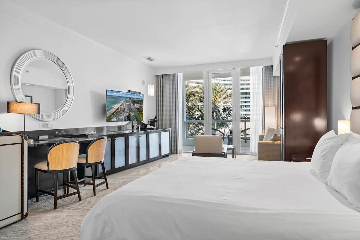 Hotelkamer met een groot bed, een bureau met stoelen, een televisie en uitzicht op palmbomen en gebouwen.