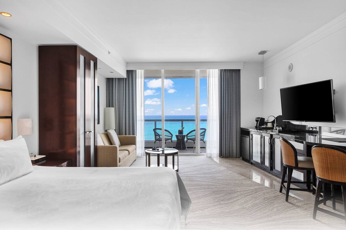 Een hotelkamer met een bed, een bureau met televisie, een balkon met uitzicht op zee, en een zithoek met stoelen.