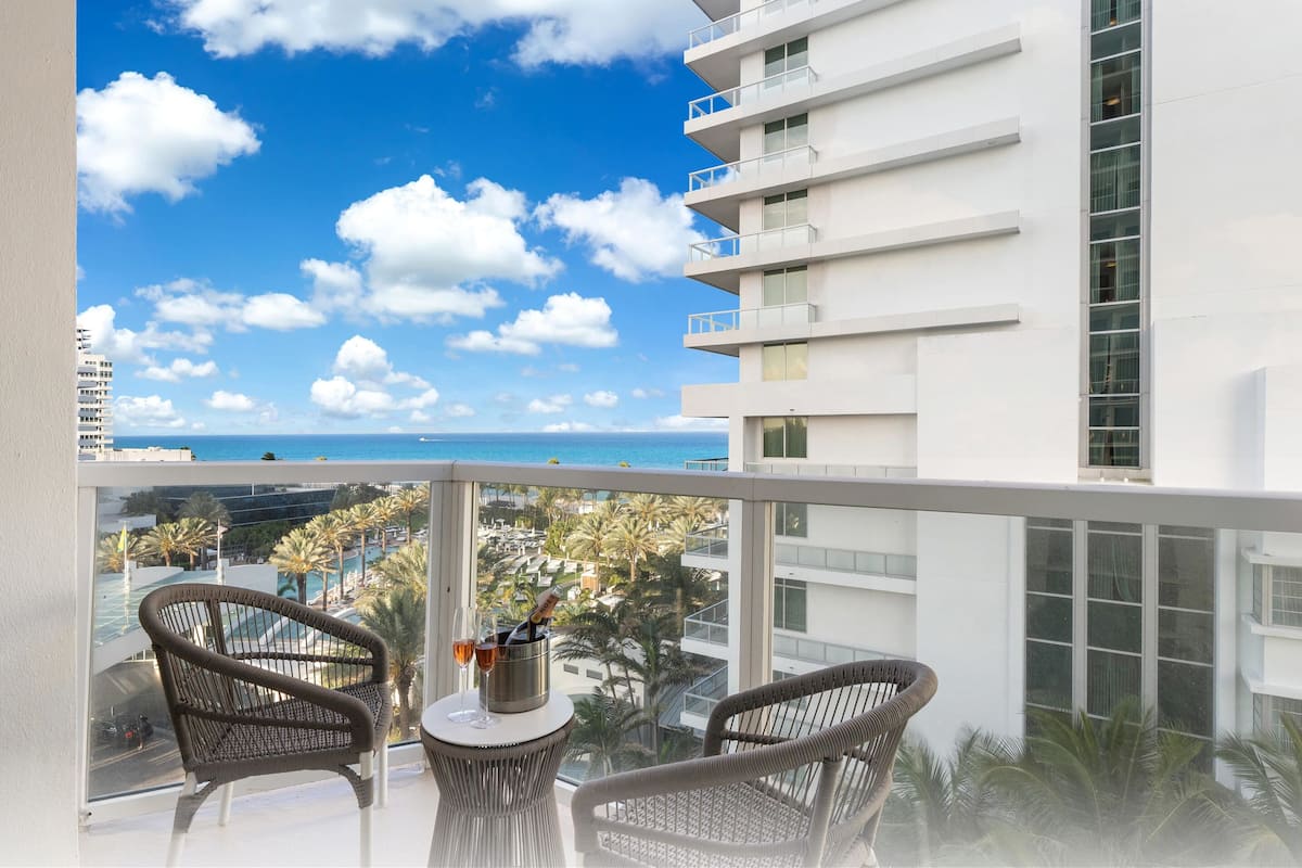 Een balkon met rieten stoelen, een tafel met een fles en glas, en uitzicht op de oceaan en palmbomen.