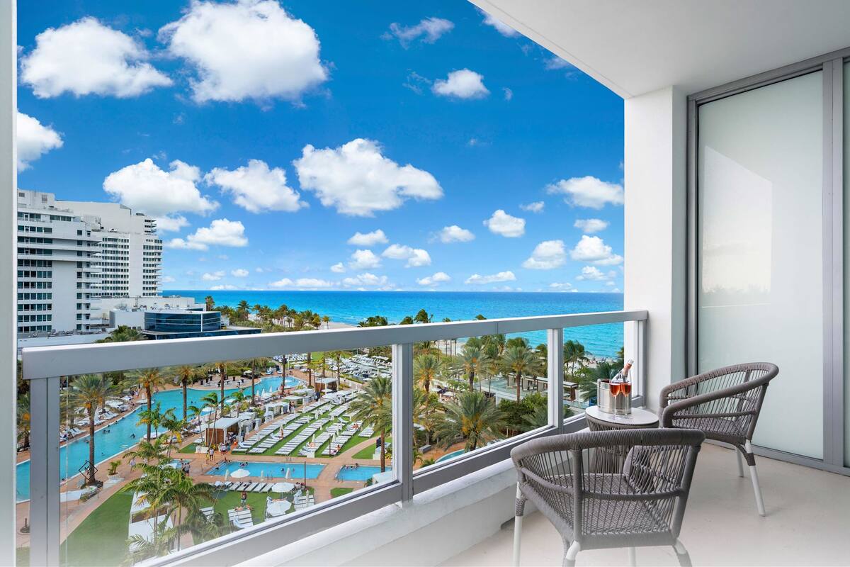 Een balkon met uitzicht op een resort zwembad, palmbomen en een strakblauwe lucht.