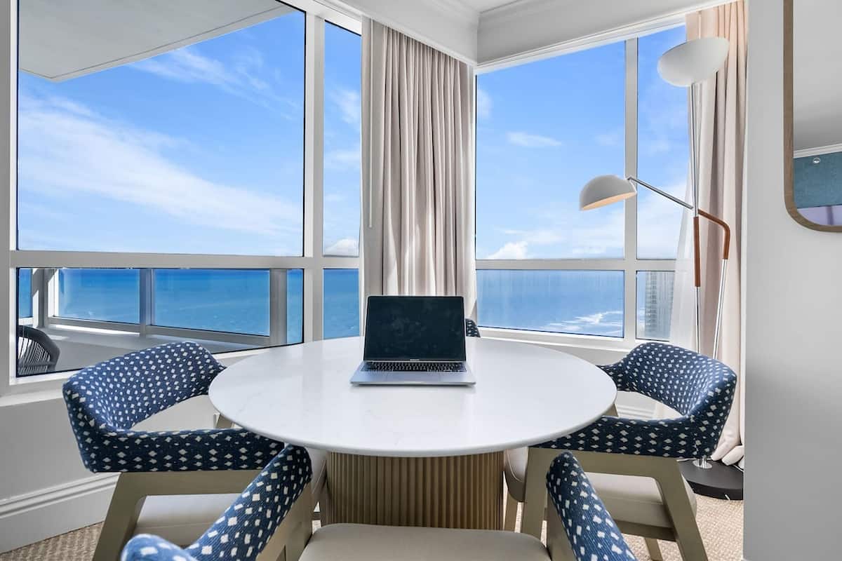 Een kamer met een ronde tafel, twee gestreept beklede stoelen, een laptop en grote ramen met uitzicht op de oceaan.