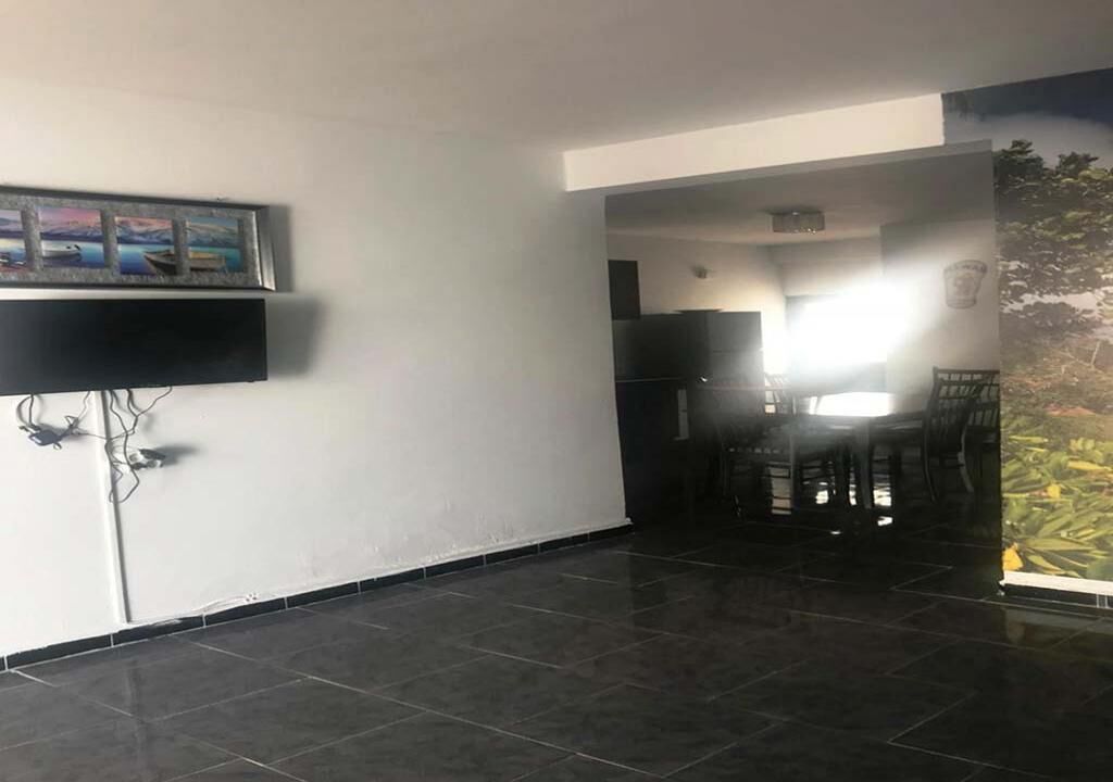 Una habitación amplia con suelo de baldosas, un televisor de pantalla plana montado en la pared y una zona de comedor con sillas y una mesa.