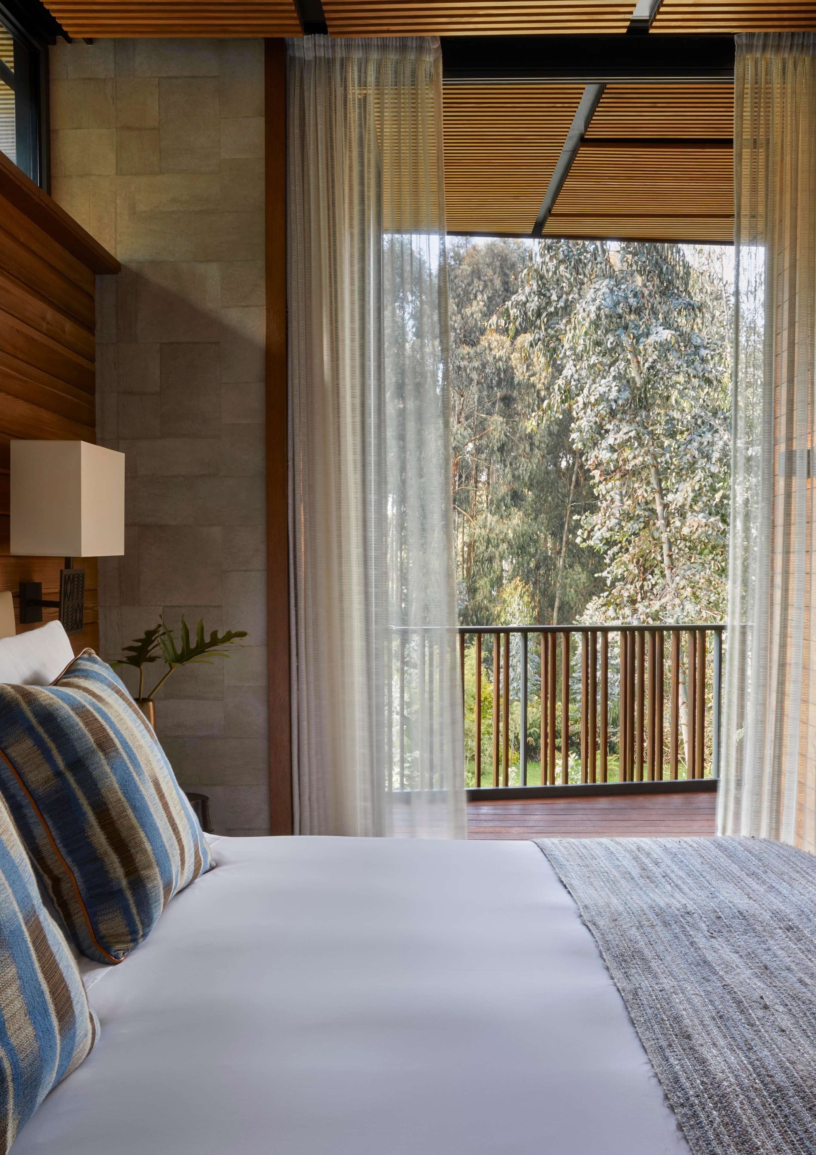 A bedroom with a large bed, a wooden headboard, a nightstand with a lamp, and a view of trees through sheer curtains.