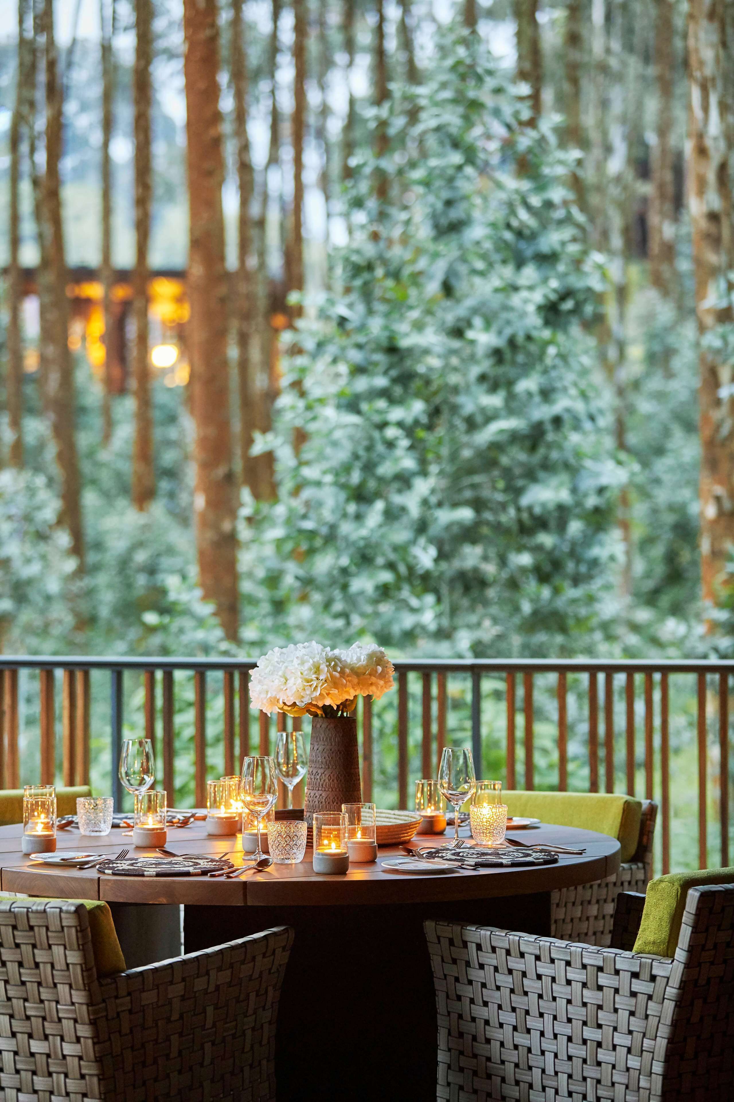 A dining table set with candles, glasses, and cutlery, surrounded by wicker chairs, with a view of a forested area.